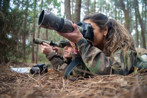 Absorbed mother and son taking pictures in forest. Family with modern cameras lying on ground, using camera and binoculars. Parenting, family, leisure concept