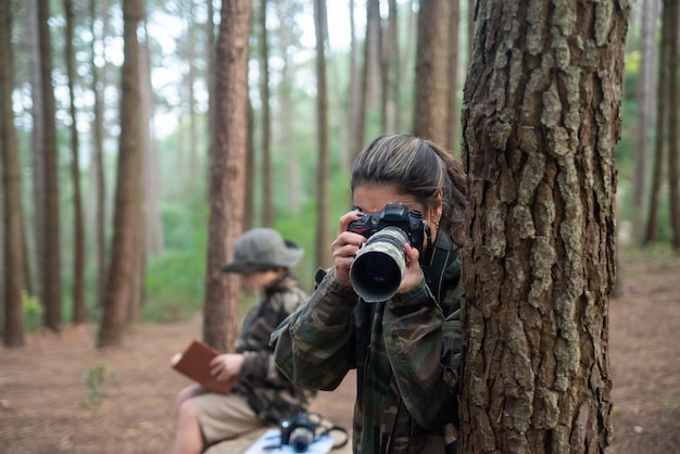 Free Photo absorbed woman  with camera in forest. mother in coat with ponytail taking pictures. blurry son in background. nature, leisure concept