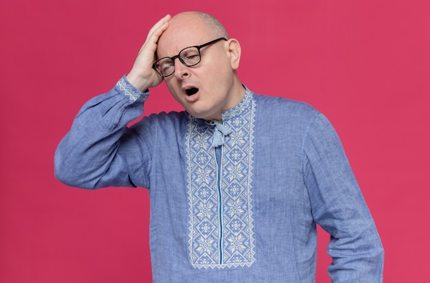 Aching adult slavic man in blue shirt wearing optical glasses putting hand on his head 