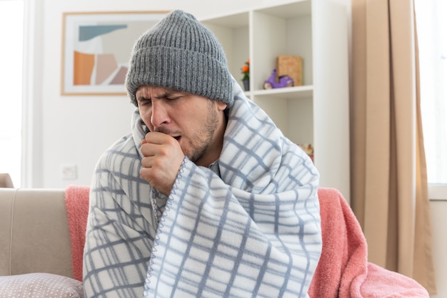 aching ill man with scarf around neck wearing winter hat wrapped in plaid coughing keeping his fist close to mouth sitting on couch at living room