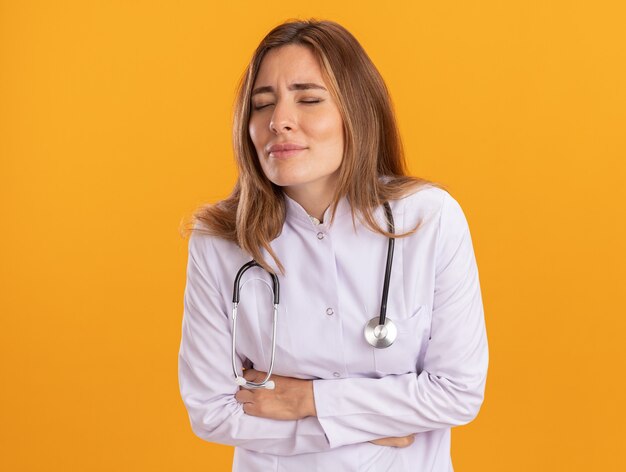 Aching with closed eyes young female doctor wearing medical robe with stethoscope grabbed aching stomach isolated on yellow wall