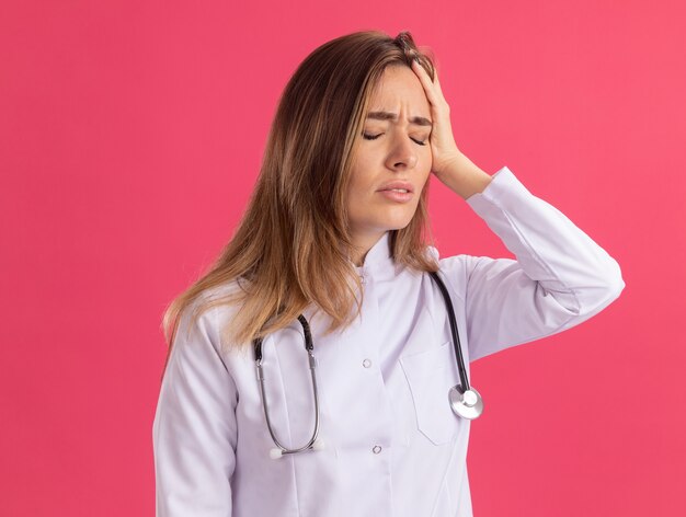 Aching with closed eyes young female doctor wearing medical robe with stethoscope putting hand on head isolated on pink wall