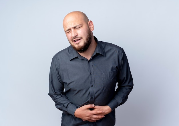 Aching young bald call center man holding his belly suffering from pain with closed eyes isolated on white  with copy space