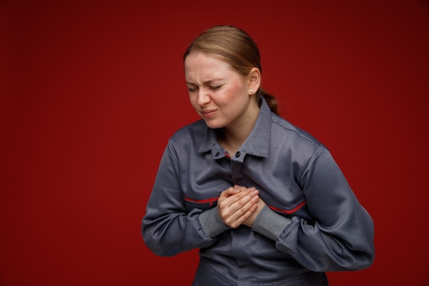 Aching young blonde female engineer wearing uniform keeping hands on chest with closed eyes 