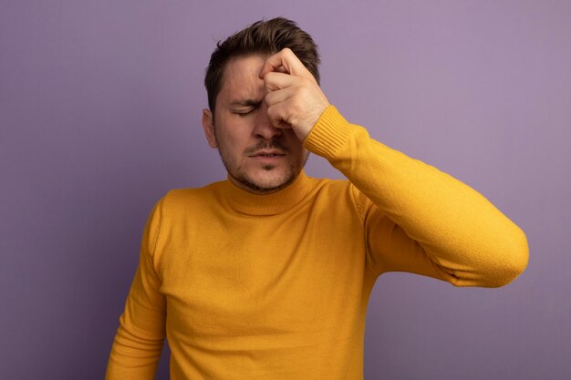 Aching young blonde handsome man keeping hand on head with closed eyes isolated on purple wall