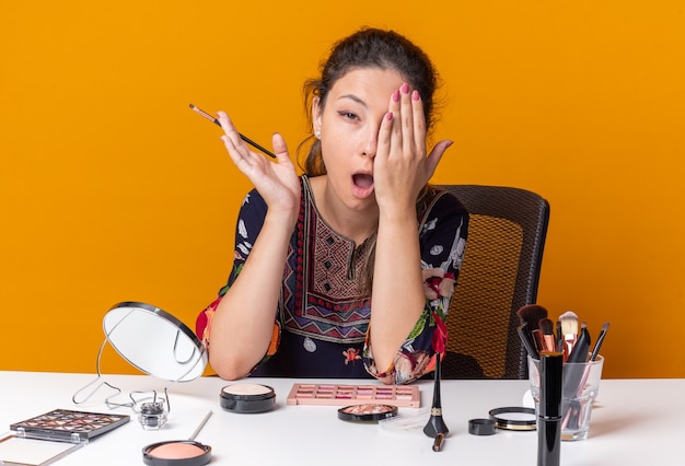 Aching young brunette girl sitting at table with makeup tools putting hand on her eye and holding makeup brush isolated on orange wall with copy space