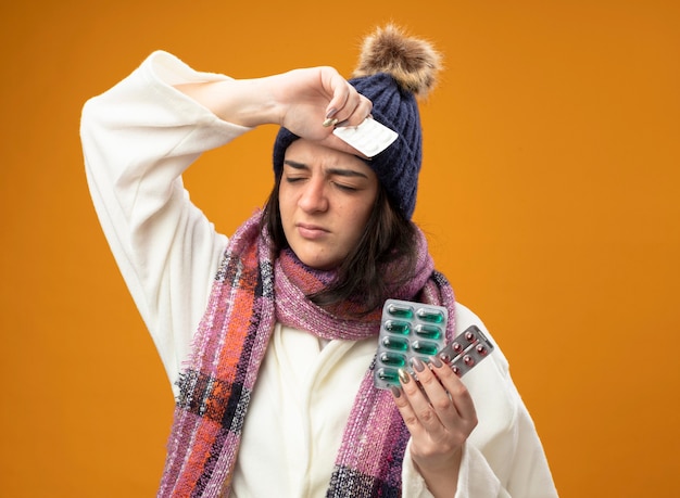 Aching young caucasian ill girl wearing robe winter hat and scarf holding packs of capsules and tablets keeping hand on head with closed eyes isolated on orange wall