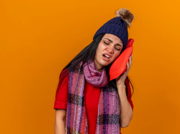 Aching young caucasian ill girl wearing winter hat and scarf touching face with hot water bag with closed eyes isolated on orange wall with copy space