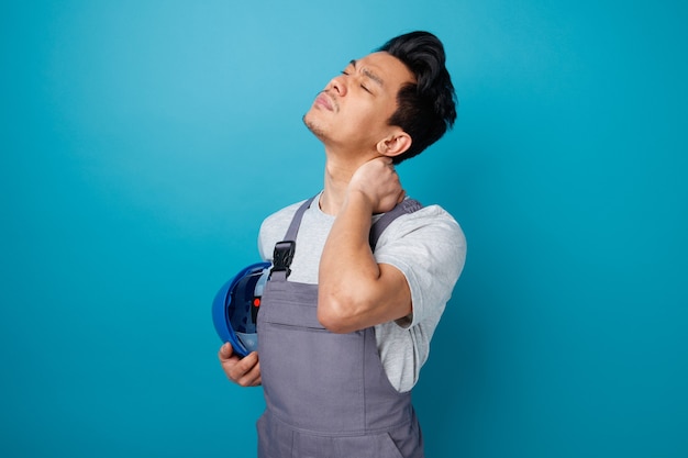 Aching young construction worker wearing safety helmet and uniform standing in profile view keeping hand on neck holding safety helmet with closed eyes 