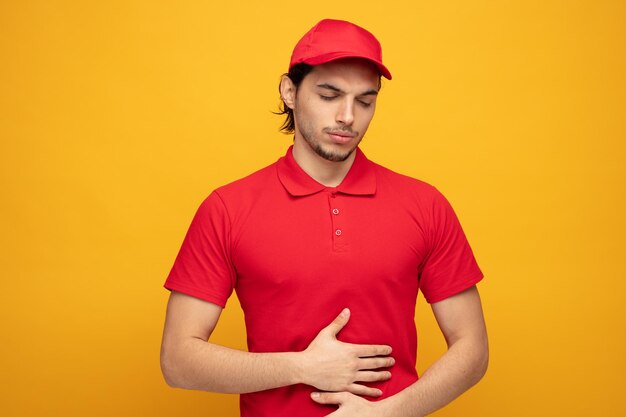 aching young delivery man wearing uniform and cap keeping hands on belly with eyes closed isolated on yellow background