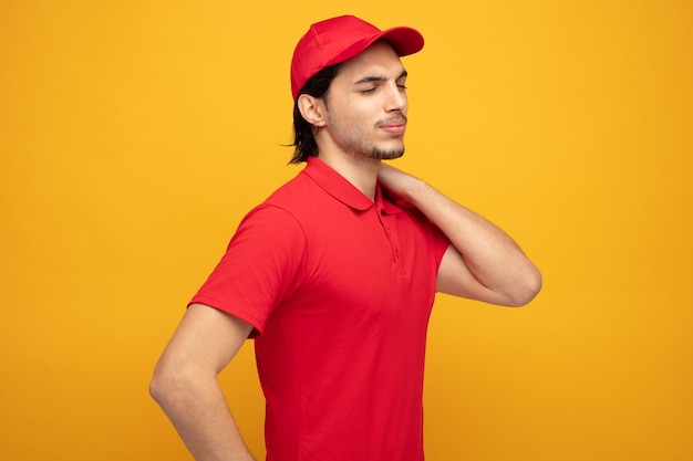 aching young delivery man wearing uniform and cap standing in profile view keeping hand on neck with eyes closed isolated on yellow background