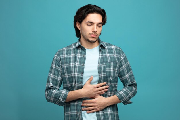 aching young handsome man keeping hands on belly with closed eyes isolated on blue background