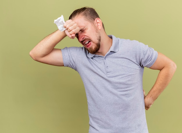 Free photo aching young handsome slavic ill man holding napkin keeping hand on waist and another one on forehead with closed eyes isolated on olive green wall