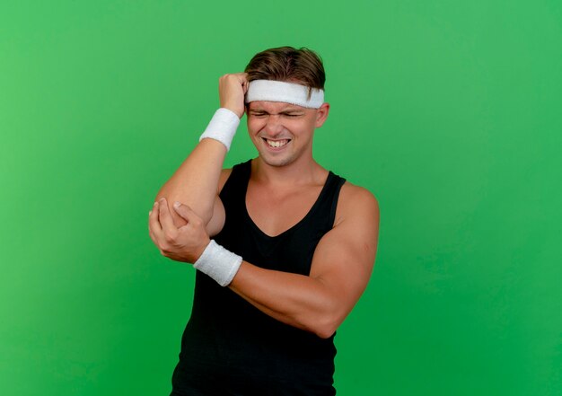 Aching young handsome sporty man wearing headband and wristbands putting hand on head and another one on elbow suffering from pain isolated on green  with copy space