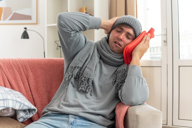 aching young ill man with scarf around neck wearing winter hat putting hand on head and holding hot water bottle sitting on couch at living room