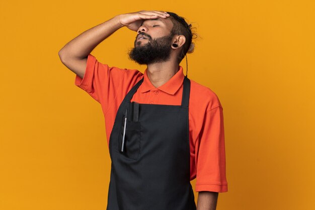 Aching young male barber wearing uniform putting hand on head with closed eyes isolated on orange wall with copy space