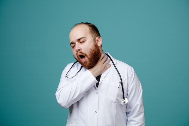 Aching young male doctor wearing medical coat and stethoscope around his neck keeping hand on neck with closed eyes having sore throat isolated on blue background