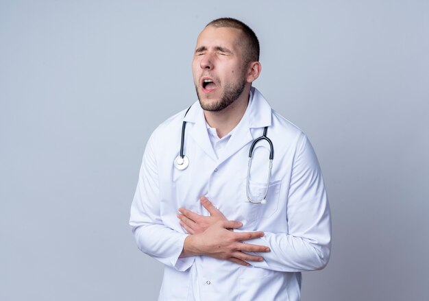 Aching young male doctor wearing medical robe and stethoscope around his neck putting hand on belly and another one on wrist with closed eyes isolated on white  with copy space