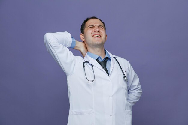 Aching young male doctor wearing medical robe and stethoscope around neck keeping hands on back and behind neck with closed eyes isolated on purple background