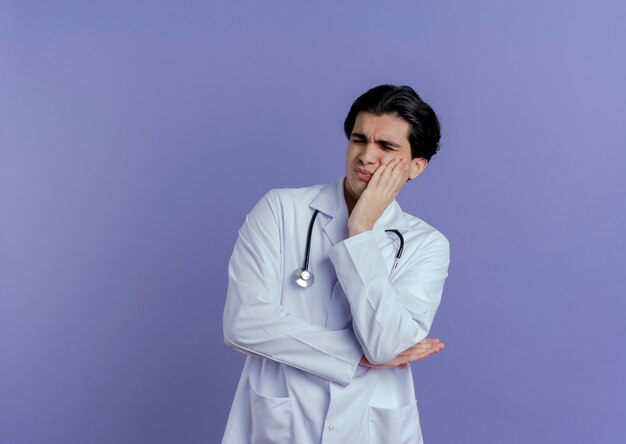 Aching young male doctor wearing medical robe and stethoscope keeping hand on cheek with closed eyes suffering from toothache isolated