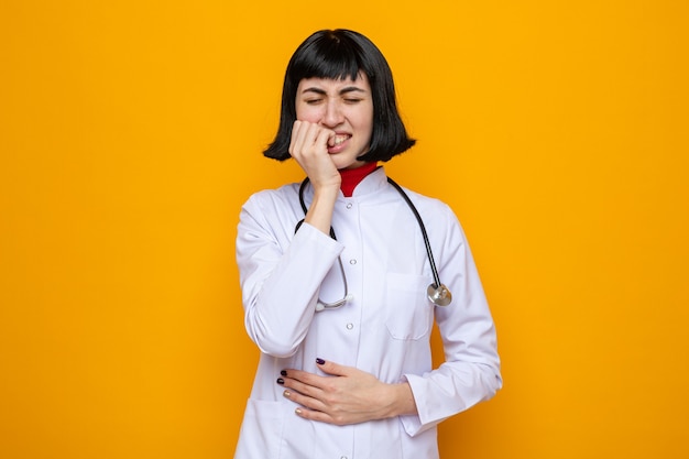 Aching young pretty caucasian girl in doctor uniform with stethoscope putting hand on belly and biting nails