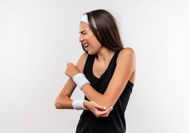 Aching young pretty sporty girl wearing headband and wristband putting hand on arm with closed eyes on white wall with copy space