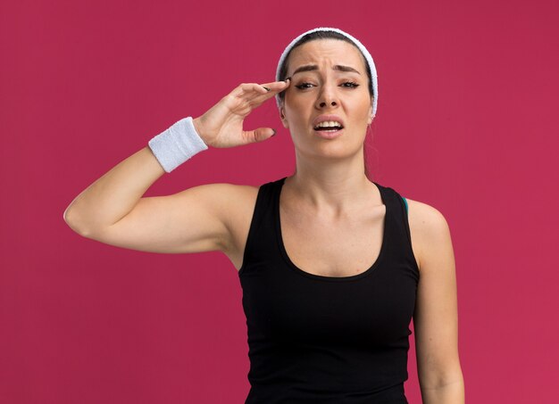 Aching young pretty sporty woman wearing headband and wristbands touching head