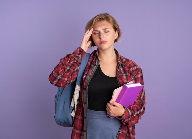 Aching young slavic student girl wearing backpack putting hand on forehead holding book and notebook 