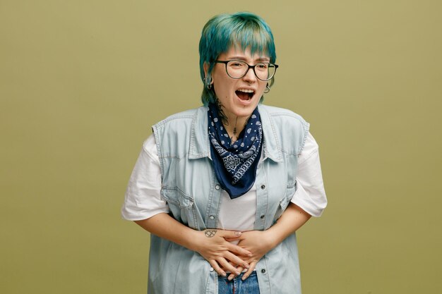 Aching young woman wearing glasses bandana on neck keeping hands on belly looking at camera isolated on olive green background