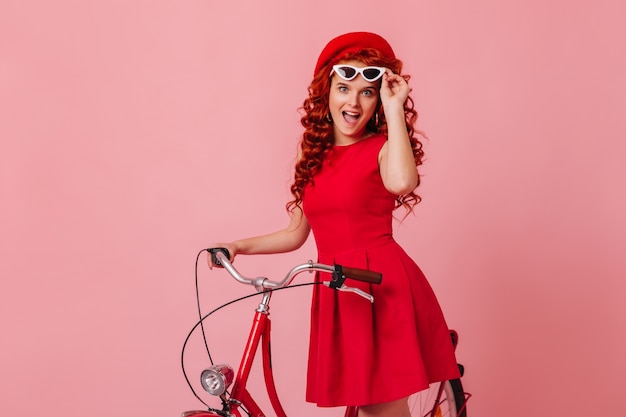 Active blue-eyed girl with curly hair takes off her glasses and looks at camera in surprise. Portrait of curly woman in red outfit riding bike.