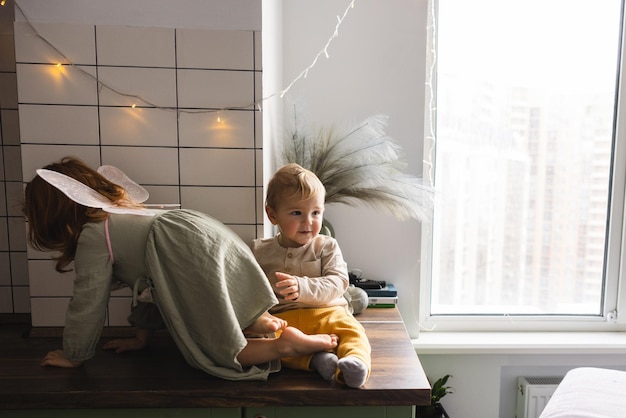 Free Photo active caucasian little redhaired girl in dress with fairhaired boy climbs on table top near window children concept