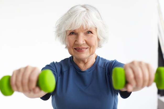 Active energetic happy elderly Caucasian female with gray hair enjoying physical exercises indoors, training at home using dumbbells, smiling broadly. Selective focus on woman's face