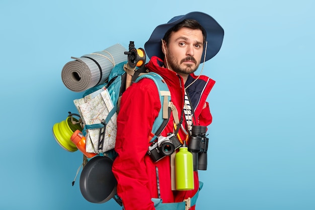 Free photo active handsome man with mustache and bristle, carries tourist rucksack on back, walks in forest, has hiking trip, wears red jacket and hat