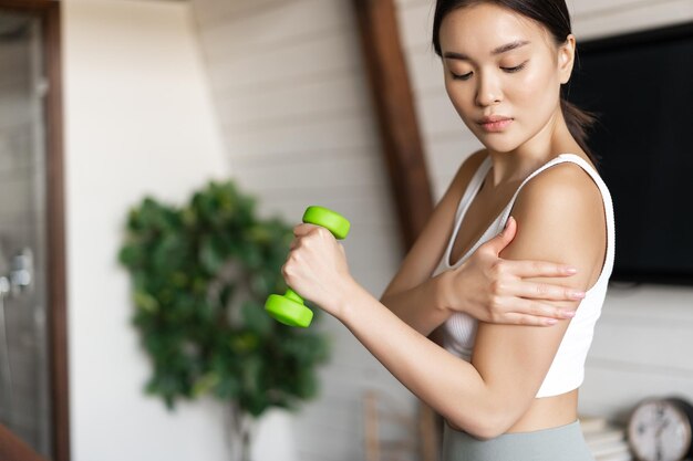 Active and healthy asian girl with fit body doing fitness exercises at home lifting dumbbells and to...