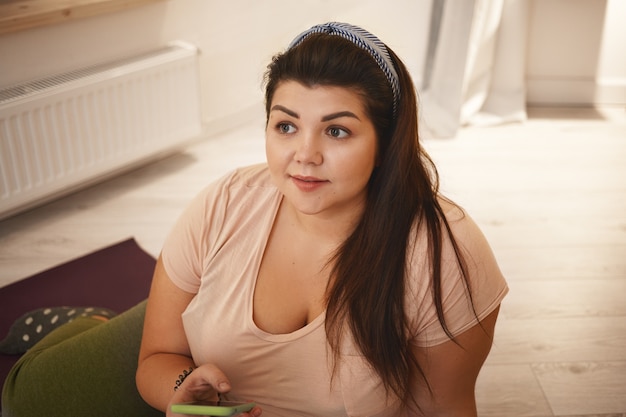 Active lifestyle, sports, health and technology. Beautiful young woman with extra pounds sitting on yoga after training, checking her weight loss results via online app on her mobile phone, smiling