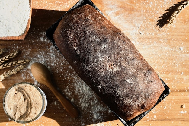 Active rye and wheat sourdough starter in a glass jar next to the ingredient flour and freshly baked whole grain bread Laid out on the table