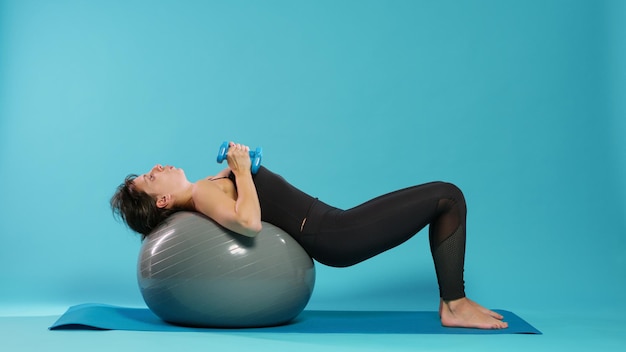 Active woman lifting dumbbells on fitness toning ball, doing physical exercise to train muscles in studio. Young adult using workout equipment and weights to practice sport and wellbeing.