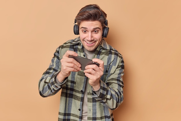 Addicted man plays video games holds smartphone horizontally tries to pass difficult level wears stereo headphones on ears dressed in checkered shirt isolated over beige background Technology