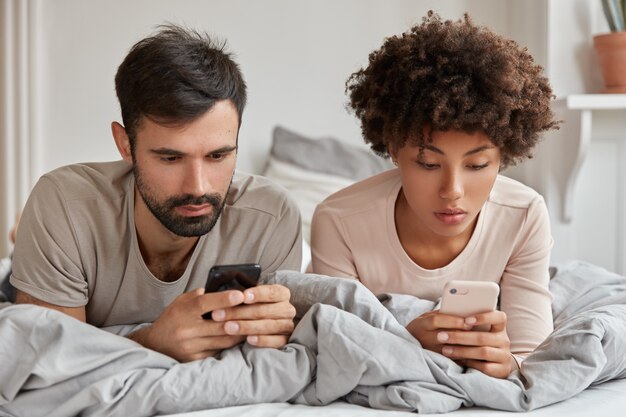 Addicted multiethnic woman and man search information in network, use modern cell phones, watch video or read weblog, have rest in bed, use wireless internet. Modern technologies and addiction