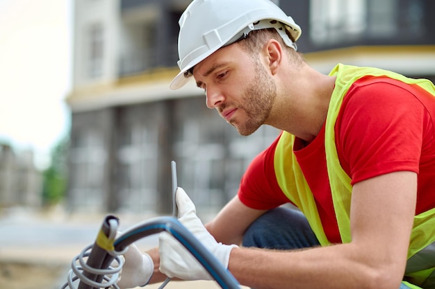 Free Photo adjustment. young adult man in protective helmet gloves and bright vest crouching looking at tool sideways to camera at construction site