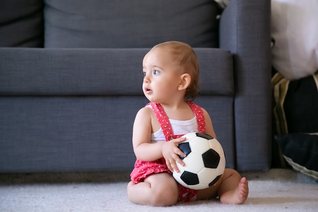 Adorable baby girl holding soccer ball, sitting on carpet barefoot and looking away. Cute infant in red dungarees shorts playing at home alone. Holiday, weekend and childhood concept