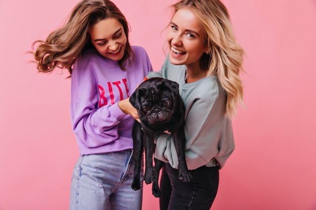 Free photo adorable blonde girl posing with her sister and french bulldog. smiling young ladies having fun with their pet.