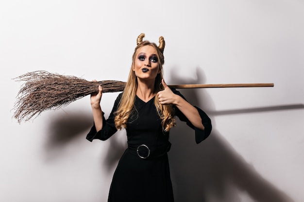 Adorable blonde witch enjoying halloween photoshoot. Elegant vampire girl in long dress holding broom.