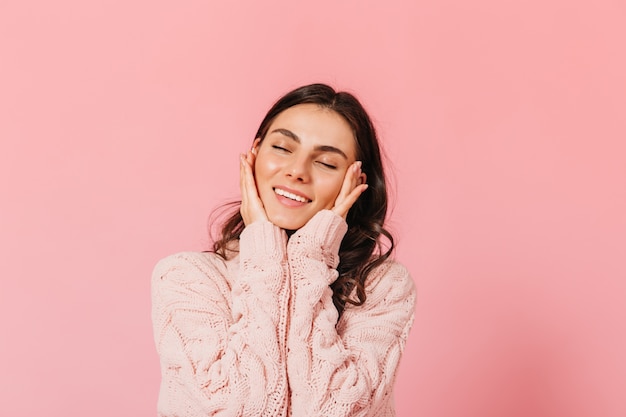 Adorable brunette woman smiles tenderly with her eyes closed. Lady in warm sweater posing in pink studio.