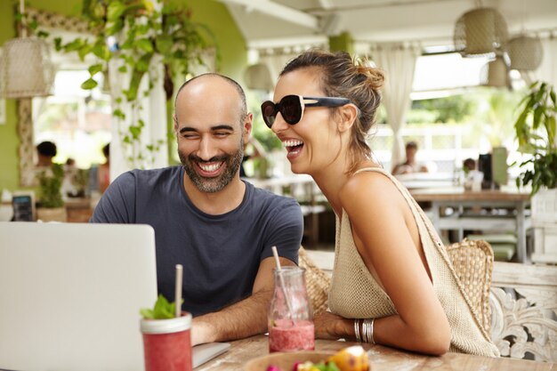 Adorable couple laughing out loud while sitting at outdoor cafe with their modern laptop and watching movies online, using free wi-fi.