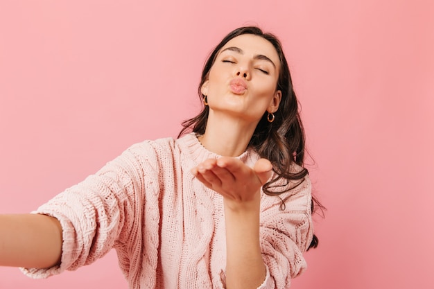 Adorable dark-haired lady in knitted sweater sends kiss and makes selfie on pink background.