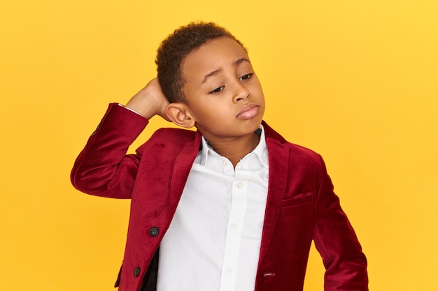 Free photo adorable dark skinned little boy pouting lips and holding hand on back of his neck, having frustrated confused facial expression, posing isolated.