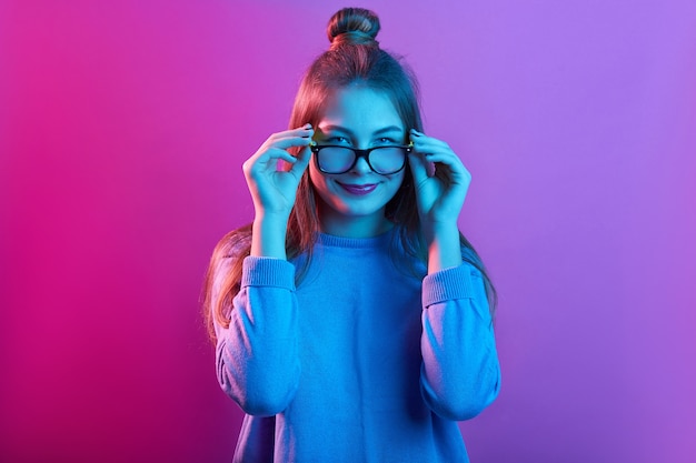 Adorable female touching frames of glasses, smiling delighted and looking at camera against pink neon wall