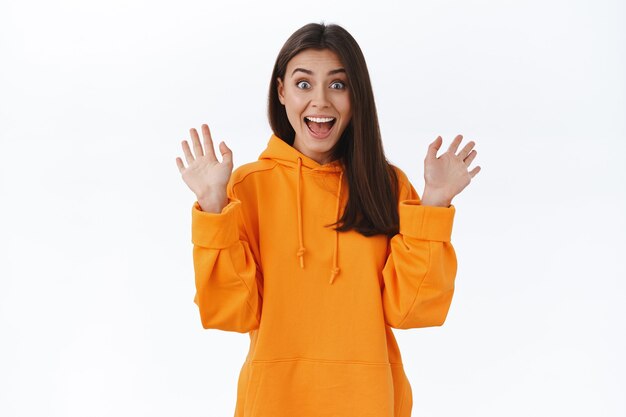 Adorable happy and surprised brunette girl seeing her friend unexpected meeting, raising hands up waving hello and scream cheerful from happiness and positive emotions, white wall