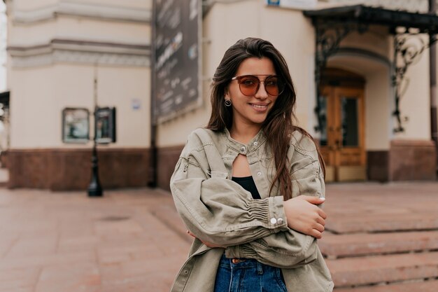 Adorable lady with long dark hair wearing denim jacket posing over old buildings in the city centre